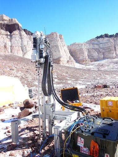 Icebreaker on a clear day in Atnarctica. The drill stands prominent in the foreground with mountains in the back beyond a rocky desert terrain with patches of ice and snow. Various equipment is around the site and part of a tent can be seen to the left.