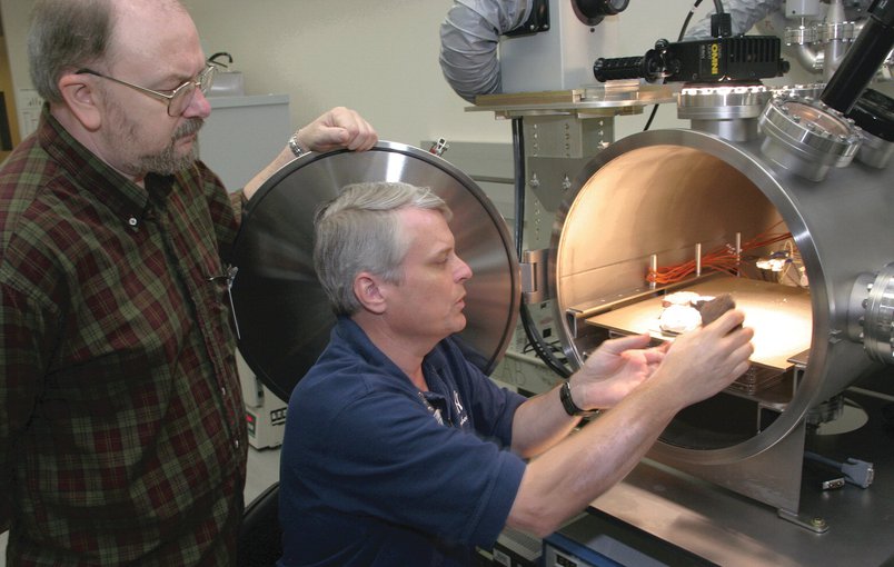 Nicholson stands to the far left in a brown check shirt holding the door to the chamber open. Schuerger crouches between Nicholson and the chamber as he places a sample in the open compartment. The chamber is a glossy, brushed metal cylinder on its side.