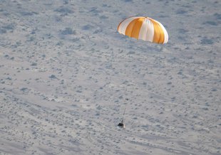 A sample return capsule is suspended from an orange and white striped parachute above a desert landscape with patchy scrub visible.
