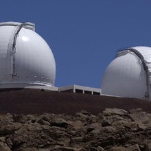 The two white domes of the Keck Observatory are seen in the daytime with a clear blue sky. The perspective is looking up toward the domes, which poke up above a rocky outcrop in the foreground.