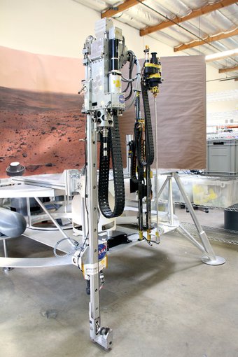 The drill stands on the concrete floor in a laboratory. A picture of the Mars surface is displayed behind it. The image is centered on the drill portion of the platform, which stands upright like a tall post. A NASA sticker is visible on the body.