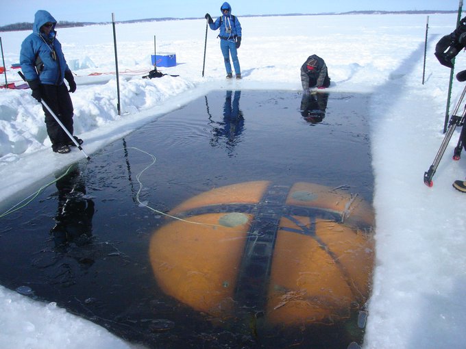 The yellow dome of ENDURANCE can be seen submerged in water, visible through a large rectangle cut through the ice of a snow-covered lake. Two scientist stand and one kneels at the end of the hole in the ice to observe. A tree line is visible at distance.